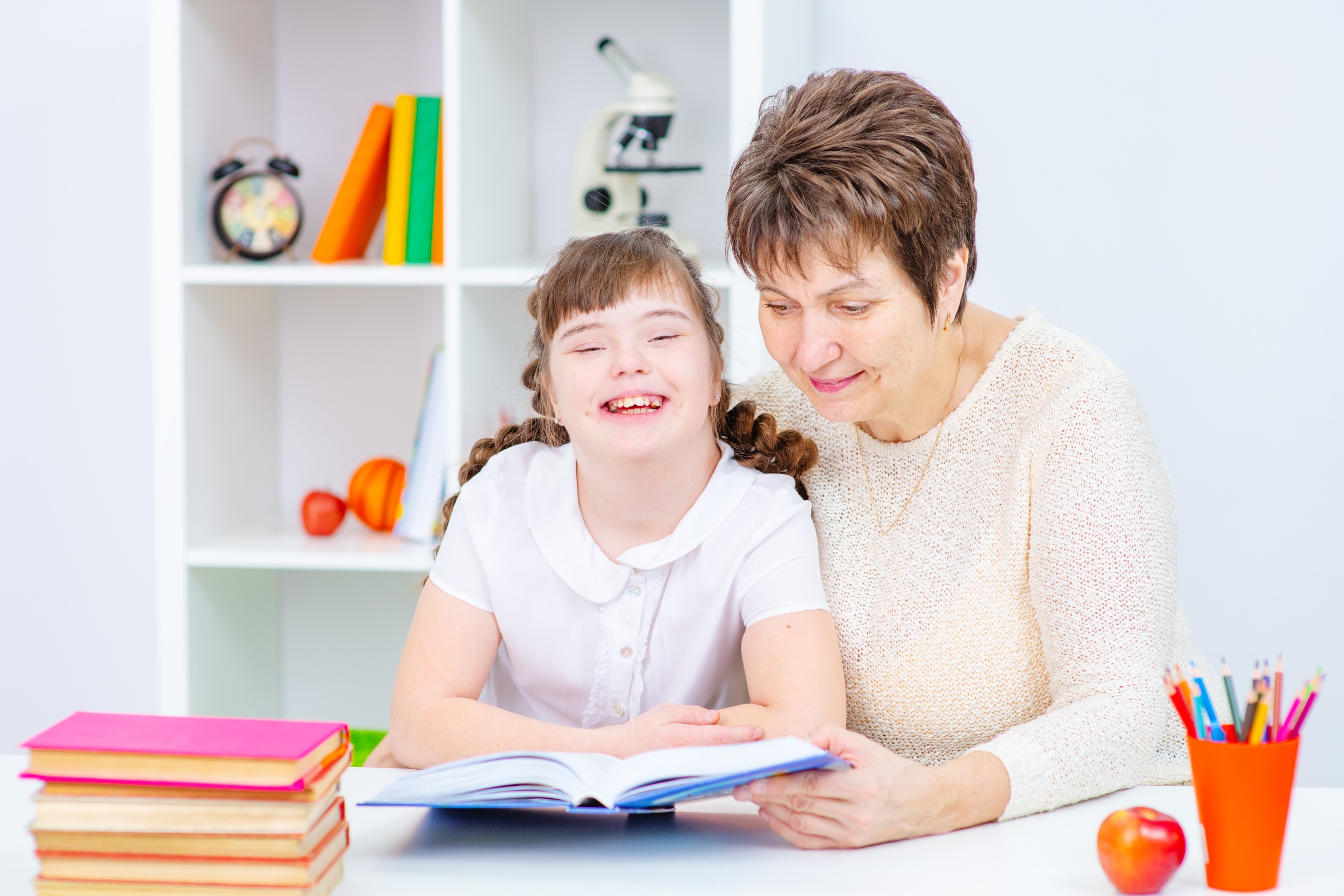 A girl with down syndrome sits at a desk in the classroom next to a tutor and an open book. The disabled child and the mother study together at home. Help for children with disabilities; Shutterstock ID 1850917765; purchase_order: Limited 3/7/22; job: ; client: ; other: