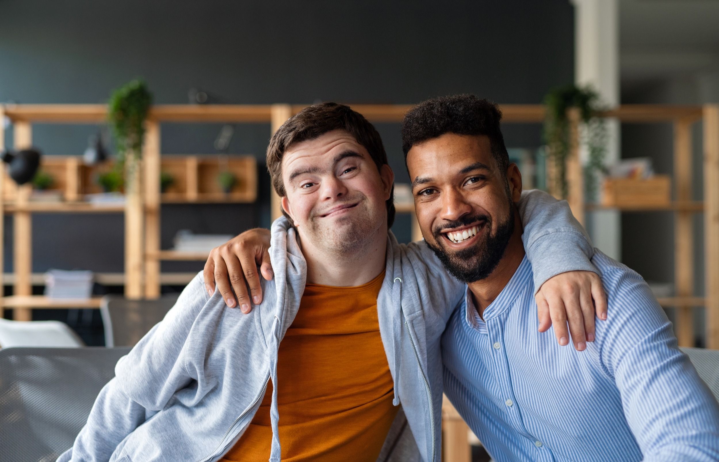 Young man with Down syndrome and his tutor with arms around looking at camera indoors at school.